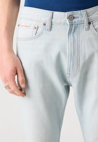 Blugi din denim de culoare bleu deschis, cu un design clasic cu cinci buzunare, nituri din cupru și o mică etichetă roșie pe buzunarul din față.