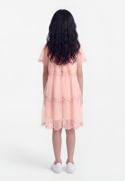 Pikkade tumedate juustega tüdruk, kes kannab heleroosat kihilist kleiti, mille äärtes on tähemustrid, ja valgeid tossusid, seisab seljaga vaataja poole.