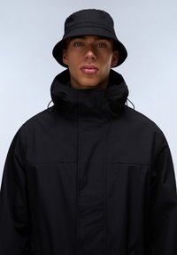Svart vattentålig jacka med hög krage och justerbar huva, i kombination med en matchande svart bucket hat. Slät textur och minimalistisk design.