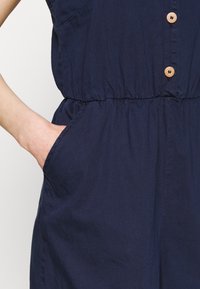 Marineblauer Jumpsuit aus weichem Stoff, mit einem gerafften Bund, Holzknöpfen und seitlichen Taschen für zusätzliche Funktionalität.