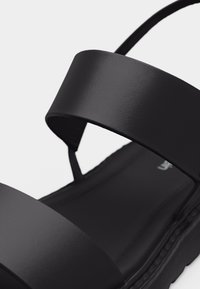 Μαύρο σανδάλι με δύο φαρδιές λουριά στο πάνω μέρος του ποδιού και επίπεδη σόλα, εμφανιζόμενο σε κοντινή λήψη πάνω σε λευκό φόντο.