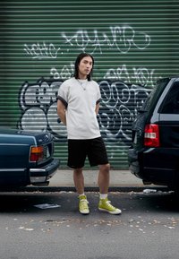 Gray short-sleeve shirt with textured hem, black shorts, and bright yellow high-top sneakers. Urban background with graffiti and parked cars.