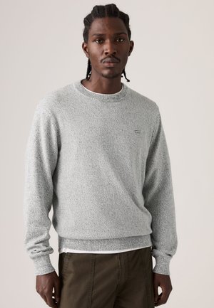 Jeune homme aux cheveux tressés portant un pull gris clair texturé à col rond et un pantalon olive foncé sur un fond uni.