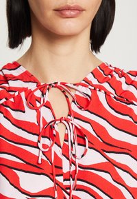 Žena v zebra pruhované blůze v červené, bílé a černé barvě s dvěma uvázanými mašlemi na výstřihu, odhalující rty, bradu a krátké tmavé vlasy.