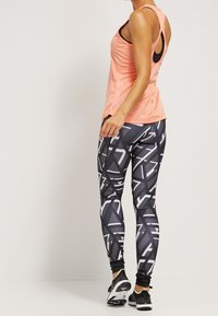  ferskenfarvet ærmeløs tanktop med scoop-hals, parret med sorte leggings med et hvidt geometrisk mønster. Smoother tekstur, atletisk pasform.