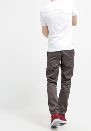 Persona con camisa blanca de manga corta, pantalones grises y zapatillas rojas, de pie con la espalda hacia el espectador.