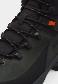 Gros plan sur une chaussure de randonnée vert foncé et noire avec des lacets noirs à pois orange passés dans des œillets en métal.
