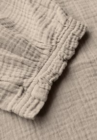 Hellbeiges, strukturiertes Material mit einer zerknitterten Oberfläche. Verfügt über ein gerafftes, fransiges Ärmeldetail. Weiches, atmungsaktives Material.