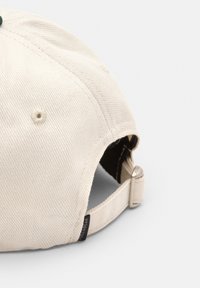 Cappello da baseball in cotone beige con una finitura testurizzata, cinturino regolabile sul retro e un piccolo etichetta nera vicino al cinturino.