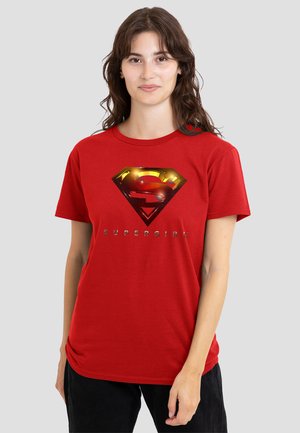 Woman with brown hair wearing a red T-shirt featuring a shiny gold and red Supergirl emblem and the word "SUPERGIRL" on a plain background.