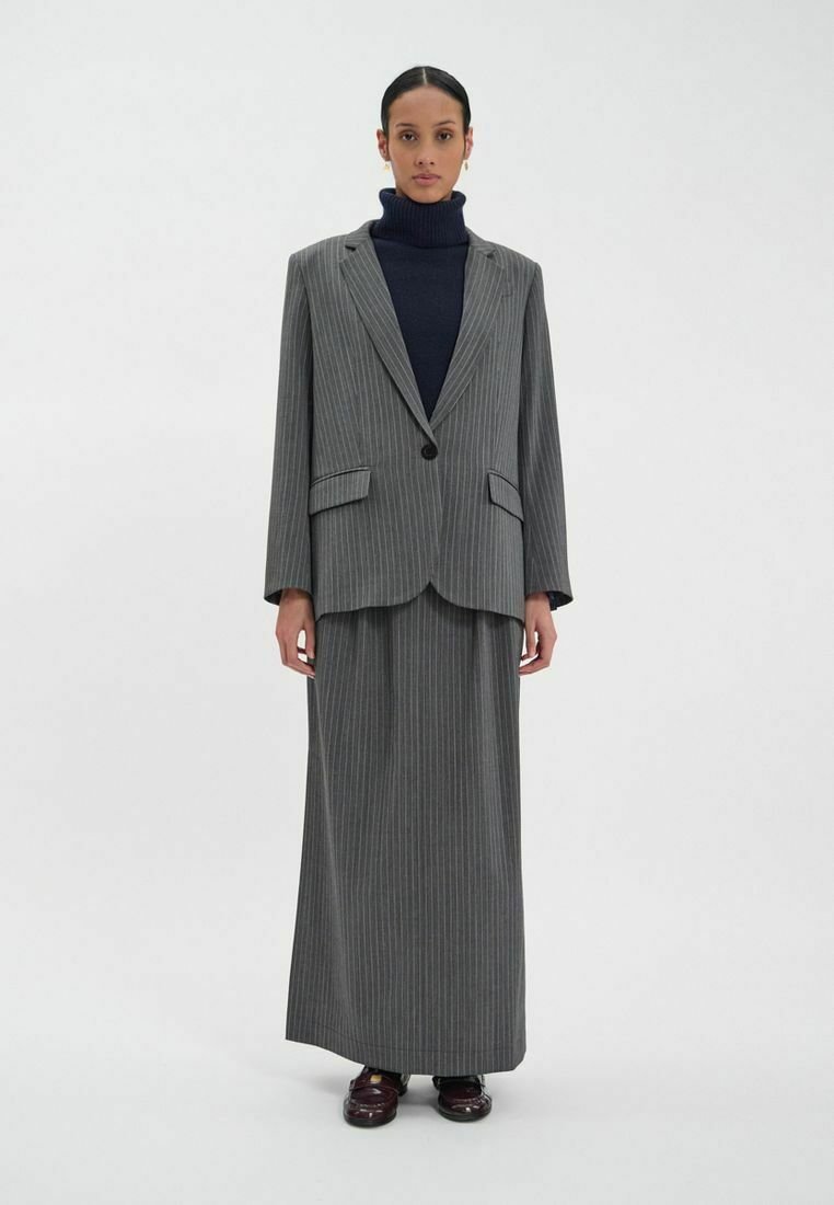 Ensemble de costume à rayures grises comprenant un blazer ajusté avec des revers en pointe, des poches à rabat et une jupe longue assortie. Porté sur un pull-collier bleu marine.