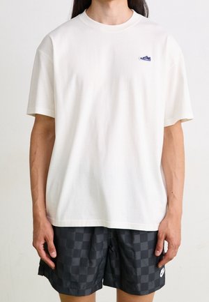 Weißes T-Shirt mit kurzen Ärmeln aus Baumwolle; verfügt über ein kleines blaues Sneaker-Logo auf der Brust; kombiniert mit dunkel karierten Shorts.