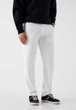 Uomo che indossa pantaloni bianchi a gamba dritta, felpa nera e scarpe da ginnastica nere con lacci bianchi, in piedi davanti a uno sfondo semplice.