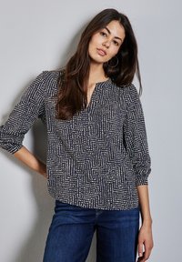 Marinblus med vitt geometriskt mönster, lättviktsmaterial, v-ringning och trekvartslånga ärmar. I kombination med mörka denimjeans.