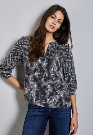 Marinebluse mit weißem geometrischem Muster, leichtem Stoff, V-Ausschnitt und Dreiviertelärmeln. Kombiniert mit dunklen Jeans.