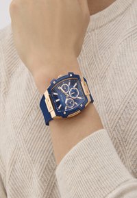 Sinine kell kuues nurkse korpuse, kuldsete aksessuaaride ja sub-dialidega sinise silikoonist rihma peal. Kandnud kät heleda sviitri vastu.