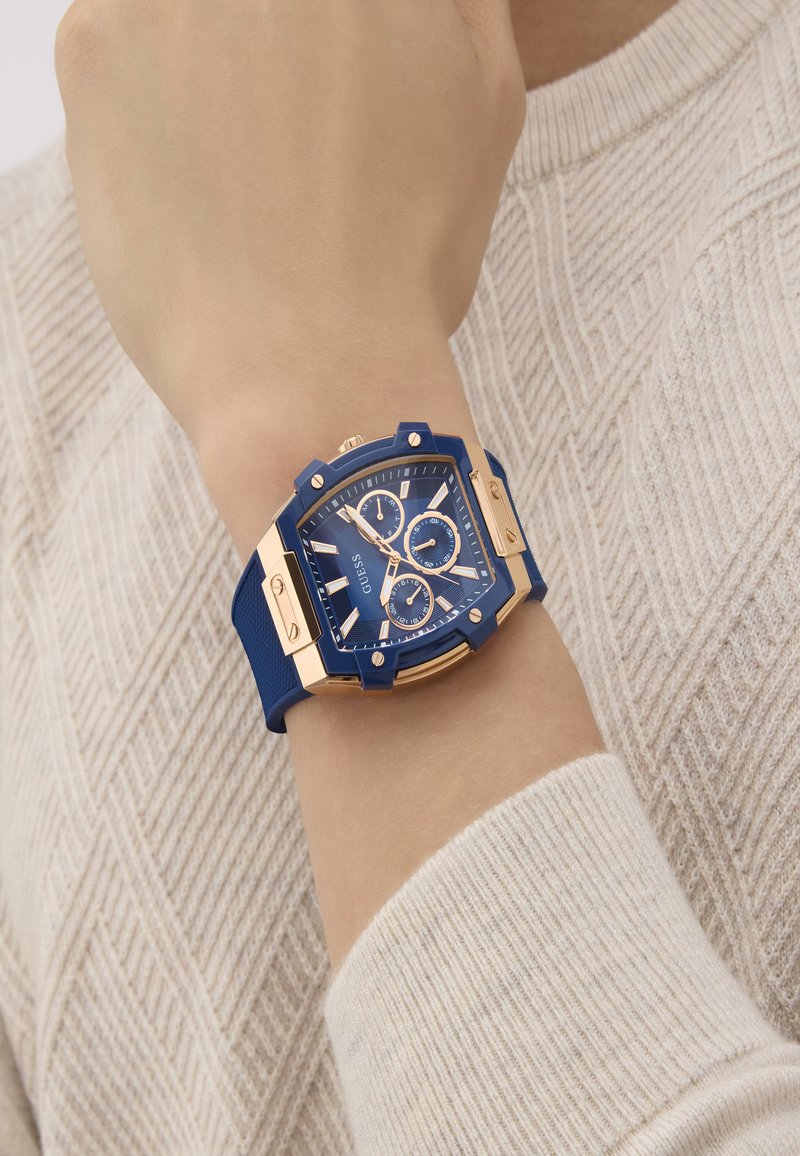 Sinine kell kuues nurkse korpuse, kuldsete aksessuaaride ja sub-dialidega sinise silikoonist rihma peal. Kandnud kät heleda sviitri vastu.