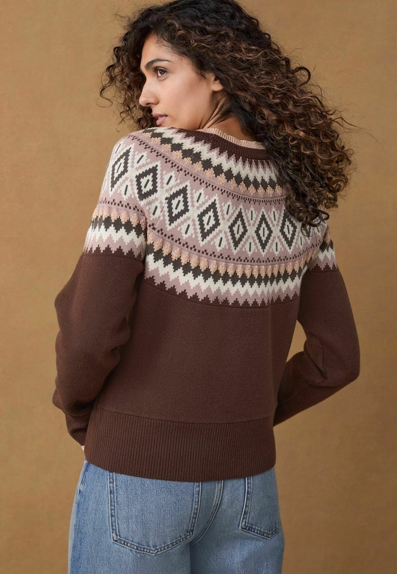 Un pull marron avec un empiècement fantaisie en crème, rose et noir avec des motifs en diamant. Poignets et ourlet côtelés. Assorti avec un jean bleu.