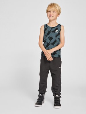 Blond boy standing and smiling, wearing a black and blue patterned sleeveless top, black pants, and black high-top sneakers.