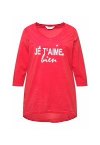 Punainen puuvillatunika, jossa on V-kaula-aukko, kolmanneksen pitkät hihat ja valkoinen teksti, joka kuuluu "JE T'AIME bien", pienillä tähden korostuksilla.