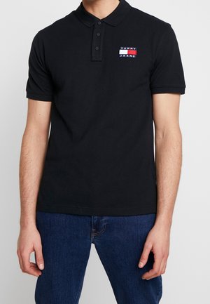 Hombre con una camiseta polo de manga corta negra con el logo "Tommy Jeans" en el pecho, combinada con vaqueros azul oscuro, de pie contra un fondo liso.