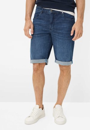 Mand iført optrukne mørkeblå denimshorts, hvide sneakers og en mørkeblå top, stående mod en ensfarvet baggrund.