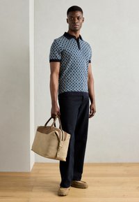 Granatowa koszulka polo w geometryczny wzór, czarne wykończenia, ciemne spodnie oraz beżowa torba podróżna ze skóry z brązowymi akcentami. Tło z drewnianej podłogi.