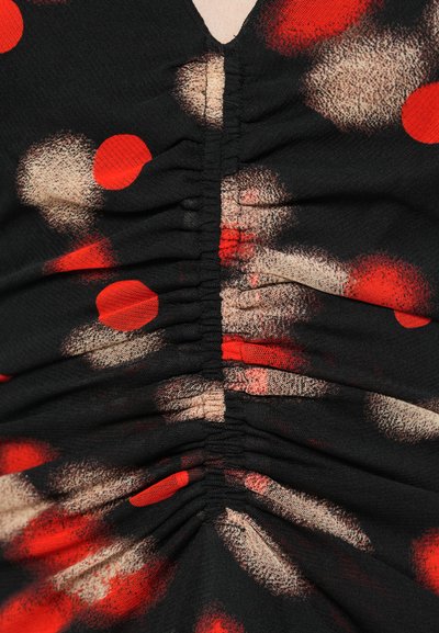 Musta kangastoppi, jossa v-pääntie ja rypytettyjä yksityiskohtia. Sisältää punaisia ja beigejä pyöreitä kuvioita, jotka luovat tekstuurisen ilmeen.