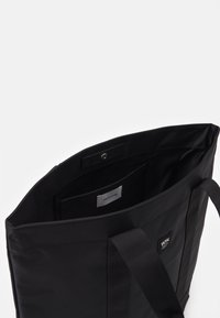 Black tote bag made of durable fabric, featuring dual handles, an internal pocket, and a snap closure, with a label visible inside.