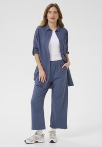 Lightweight, button-up shirt in slate blue, paired with loose, matching pants. Model wears sneakers with white socks. Casual, comfortable fit.