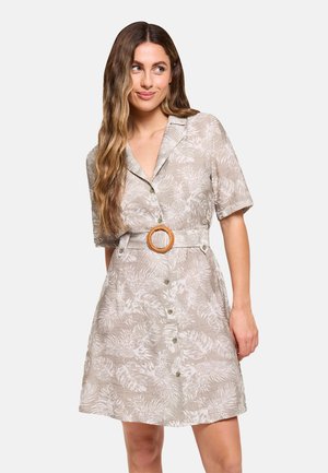 Mujer con el pelo largo y ondulado, que lleva un vestido beige de manga corta con botones, estampado floral blanco y cinturón con hebilla redonda.