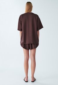 Bruin oversized overhemd met korte mouwen en bijpassende korte broek, gemaakt van zachte stof. De outfit heeft een ontspannen pasvorm en wordt gecombineerd met eenvoudige sandalen.