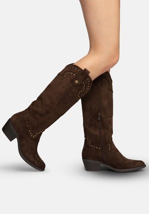 Brune ruskinds-knæhøje støvler med lave stakkede hæle, sidelåse og dekorative messingnitter, båret på bare ben mod en ensfarvet baggrund.