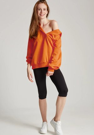 Pomarańczowy oversize'owy sweatshirt z dekoltem off-shoulder, zestawiony z czarnymi legginsami i białymi sneakersami. Gładki materiał i luźny krój.