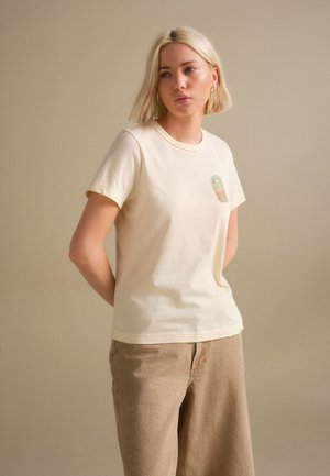 Jeune femme avec une coupe au carré blonde, portant un t-shirt crème avec un petit graphisme et un pantalon beige ample, debout devant un fond uni.
