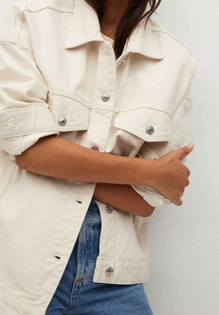 Cream-colored oversized denim jacket with chest pockets, silver snap buttons, and rolled sleeves, paired with blue denim jeans.