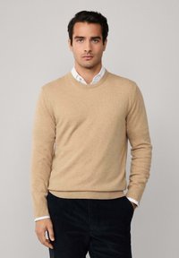 Muškarac u bež puloveru s okruglim izrezom preko bijele košulje s dugim rukavima, u crnim hlačama, stoji naslonjen na jednostavnu svijetlo sivi pozadinu.