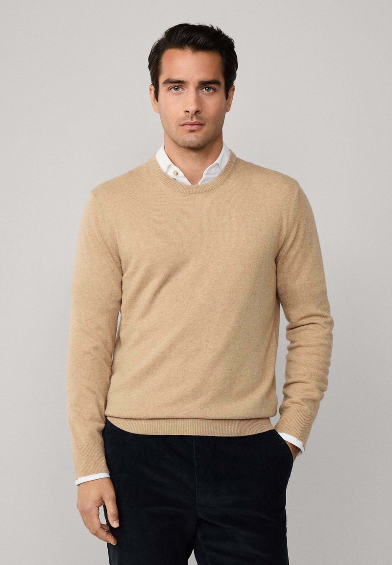 Muškarac u bež puloveru s okruglim izrezom preko bijele košulje s dugim rukavima, u crnim hlačama, stoji naslonjen na jednostavnu svijetlo sivi pozadinu.