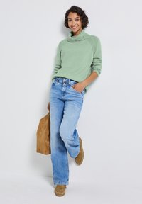 Světlazelený pletený svetr s krouceným výstřihem, zkombinovaný se světle modrými džínami. Hnědé semišové boty a béžová taška dotvářejí vzhled.