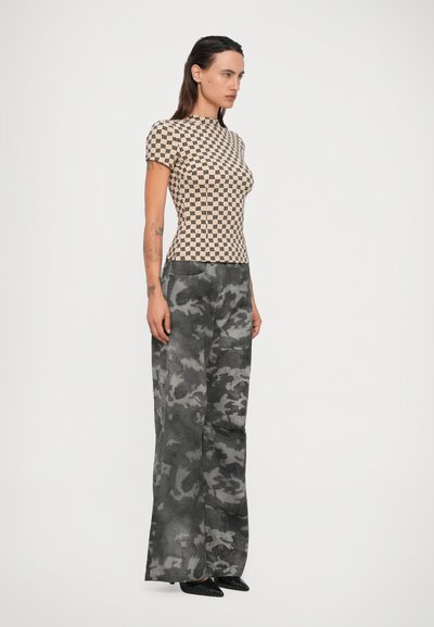 Eine Frau trägt ein beiges, gemustertes Kurzarmoberteil und lockere schwarz-graue Camouflage-Hosen. Sie steht seitlich vor einem einfarbigen Hintergrund.