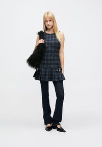 Túnica sem mangas em padrão xadrez azul-marinho e verde com bainha franzida, combinada com jeans flare azul-escuro e sapatilhas pretas, segurando uma mala de pele sintética preta.