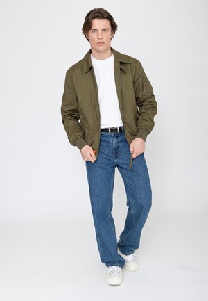 Mann mit braunen Haaren, der eine grüne Jacke, ein weißes Hemd, blaue Jeans und weiße Turnschuhe trägt, steht vor einem schlichten Hintergrund.