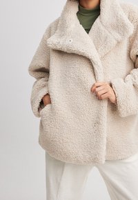 Beige textuur teddyjas met grote kraag en knopen, gedragen over een olijfgroen top en witte ribgebreide broek, handen in de zakken en de jas vasthoudend.