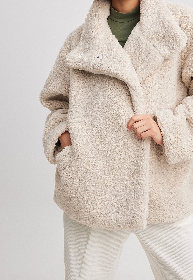 Beige textuur teddyjas met grote kraag en knopen, gedragen over een olijfgroen top en witte ribgebreide broek, handen in de zakken en de jas vasthoudend.