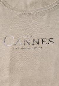 Beige bavlněné tričko s kulatým výstřihem,  s tmavě šedým nápisem "CLUB CANNES" a sloganem pod ním. Jednoduchá textura, bez vzorů nebo akcentů.