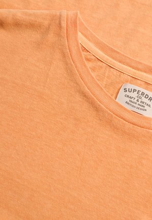 Jasnopomarańczowa koszulka z okrągłym dekoltem, z widoczną wszywką marki Superdry wewnątrz kołnierzyka.