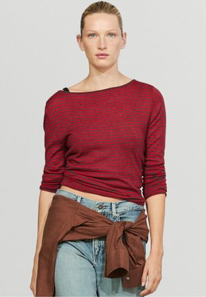 Femme aux cheveux blonds portant un haut à manches longues rayé rouge et noir et un jean bleu clair avec une veste marron nouée autour de la taille.