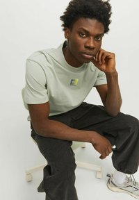 Camiseta de algodón verde claro con un logo cuadrado en azul y amarillo en el pecho, combinada con pantalones negros holgados y zapatillas grises.