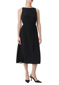 Nainen, joka on pukeutunut olkaimettomaan mustaan midi-mekkoon, jossa on ohut vyö vyötäröllä, ja mustiin teräväkärkisiin slingback-korkokenkiin, seisoo valkoista taustaa vasten.