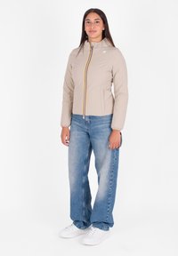 Jonge vrouw in een beige zip-up jack, losse blauwe jeans en witte sneakers, staande tegen een plain witte achtergrond.